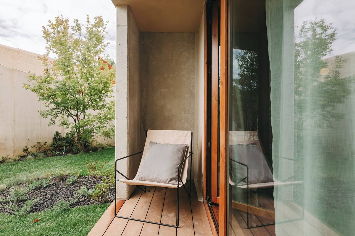 Ein Balkon mit gepolstertem Stuhl, einer Glastür und einem kleinen Garten mit Baum und Rasen.