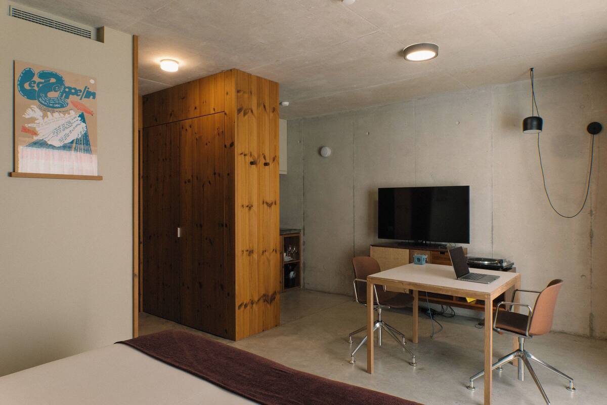 Ein modernes Zimmer mit einem Bett, einem Holzschrank, einem Schreibtisch mit einem Laptop, einem Fernseher und einem Sessel.