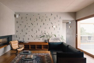 A modern living room with a black sofa, a wooden coffee table, a patterned rug, a leather armchair, and a wall with abstract designs.