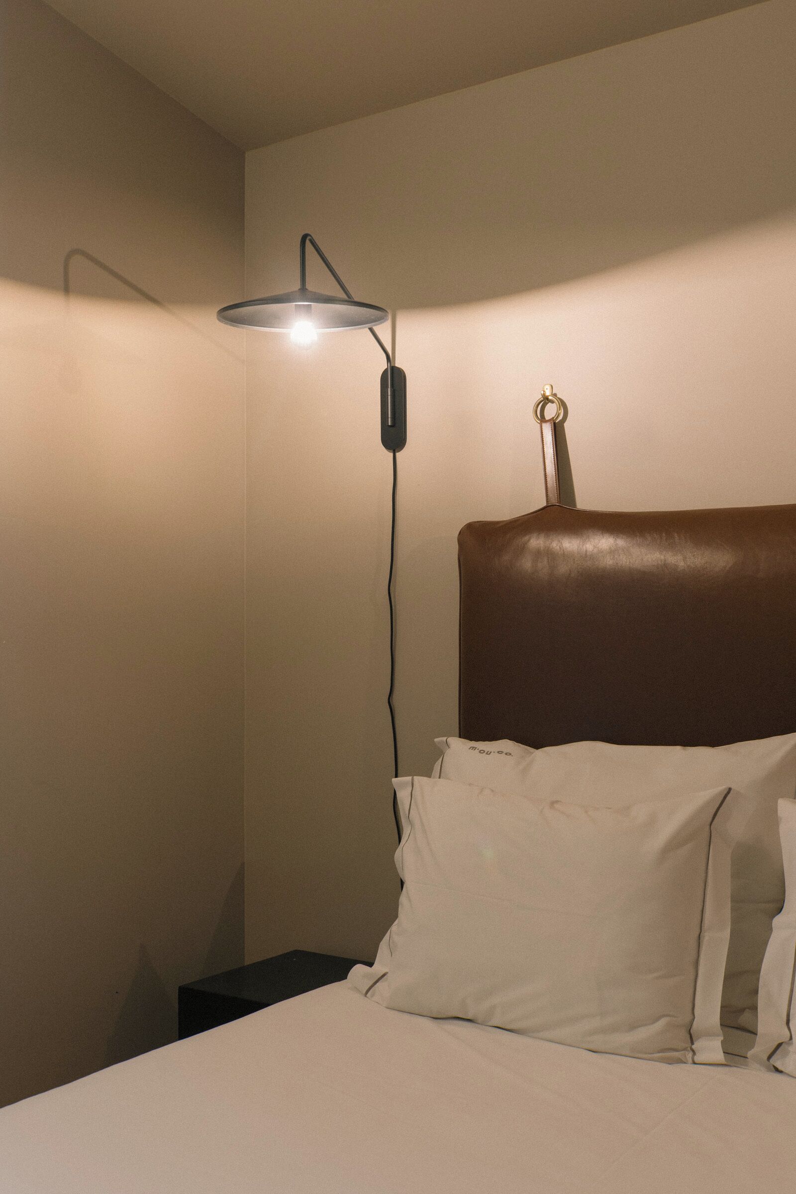 Un lit bien fait, avec du linge de lit blanc et une tête de lit marron, une table de chevet et une lampe de sol avec un pied noir et un abat-jour blanc.