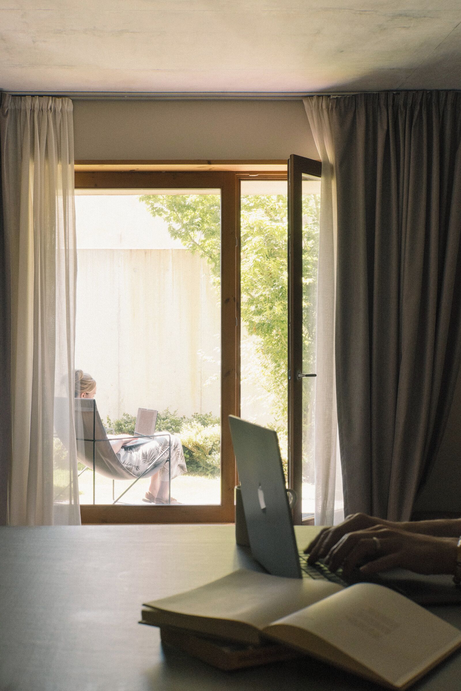A room with a laptop on a table, an open book, and a view through a window with curtains.