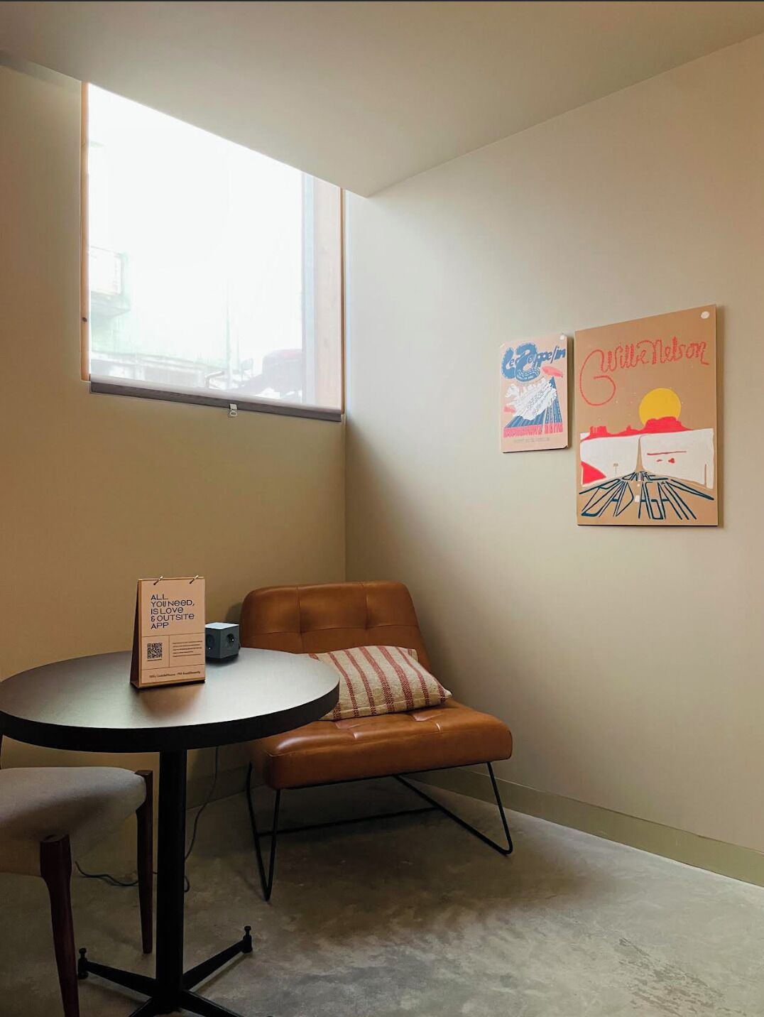 Een kamer met een raam, een bruine leren fauteuil, een klein tafeltje en twee kunstwerken in lijsten aan de muur.