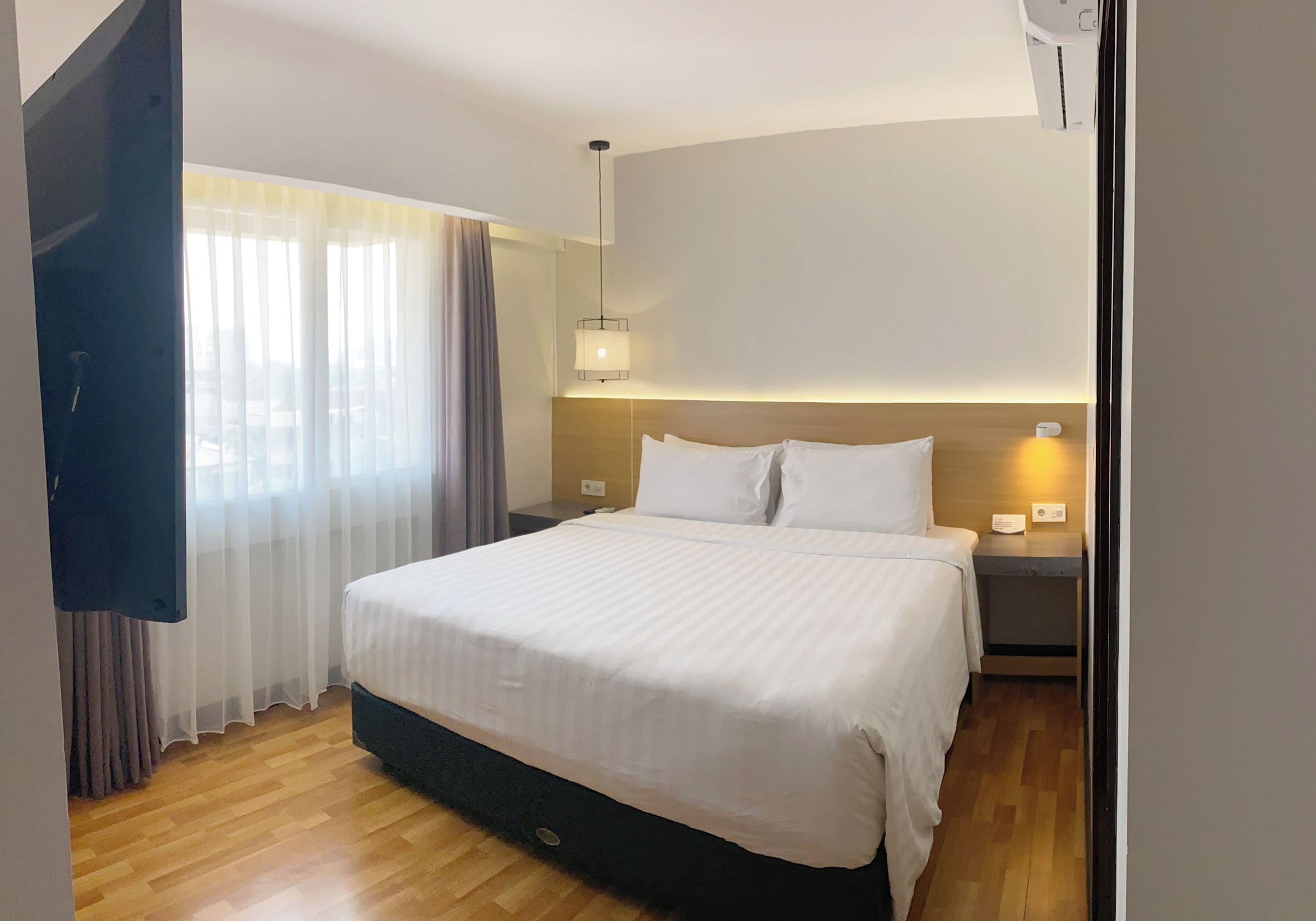 Quarto de hotel com cama, mesa de cabeceira, luminária e janela com cortinas.