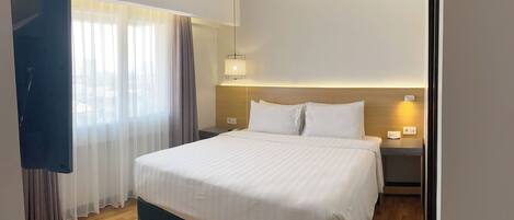 Quarto de hotel com cama, mesa de cabeceira, luminária e janela com cortinas.