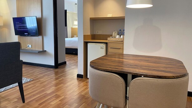 Habitación de hotel moderna con una mesa de comedor de madera, dos sillas, un televisor montado en la pared, una zona de cocina y una puerta que conduce a otra habitación.