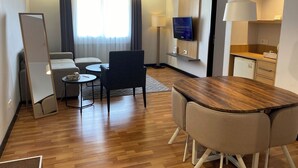 Habitación de hotel con piso de madera, un sofá, una mesa de comedor con sillas, un televisor montado en la pared y una pequeña cocina.