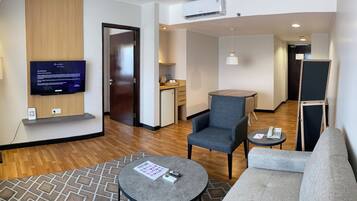 A modern hotel room with a flat-screen TV, a sofa, a chair, a small table, and a kitchenette in the background.