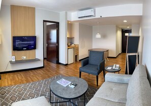 Une chambre d’hôtel moderne équipée d’un téléviseur à écran plat, d’un canapé, d’un fauteuil, d’une petite table et d’une kitchenette à l’arrière-plan.