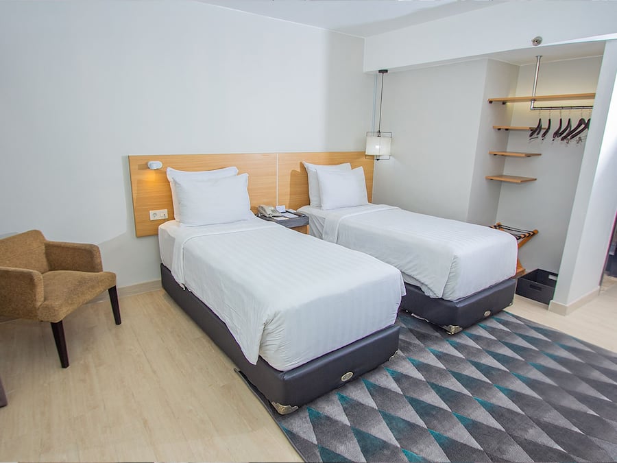 Quarto de hotel com duas camas, uma cadeira, mesa de cabeceira, abajur e guarda-roupa.