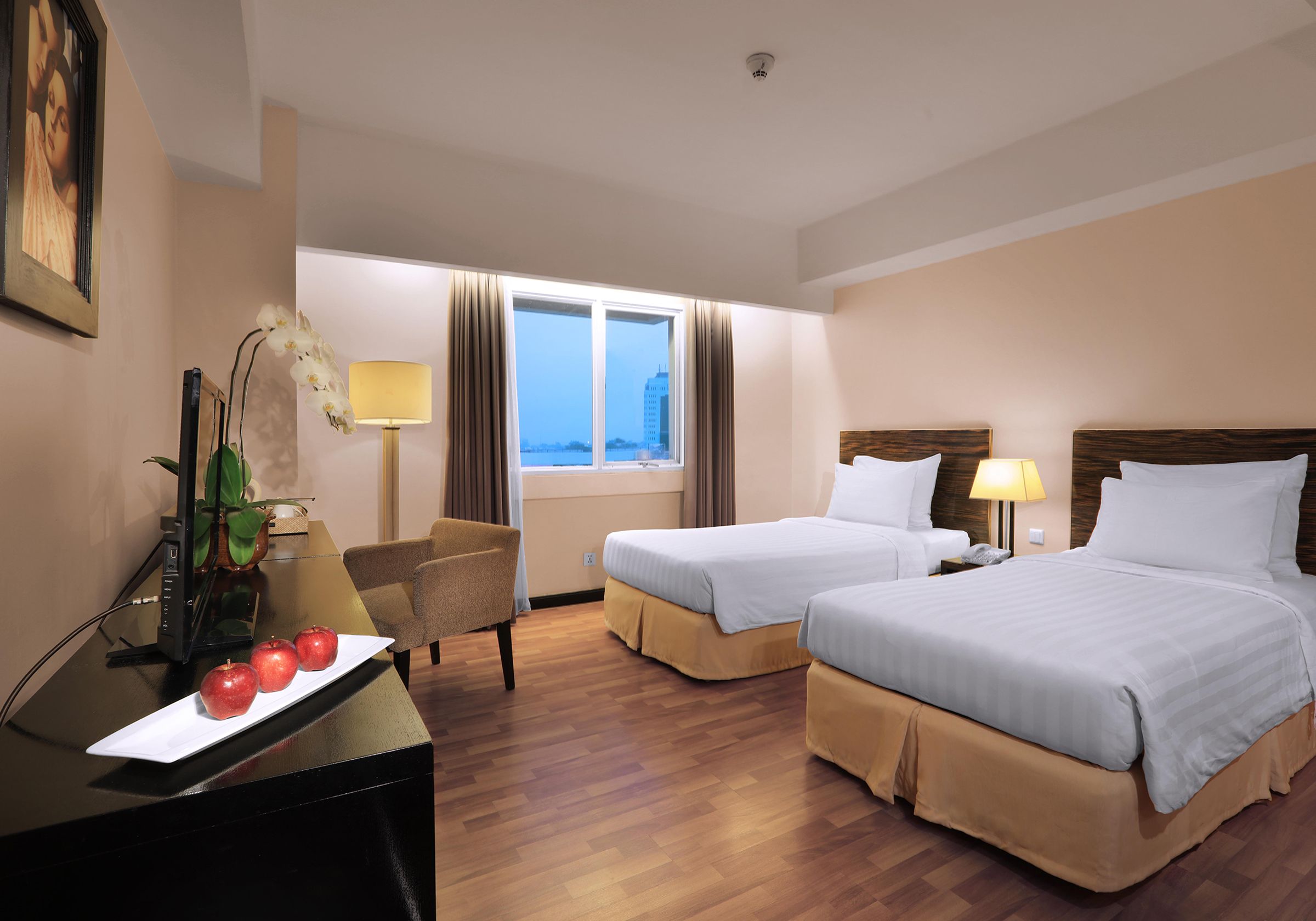 A hotel room with two beds, a desk with a computer and apples, a chair, a lamp, and a window with curtains.
