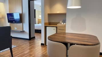 Habitación de hotel moderna con una mesa de comedor de madera, dos sillas, un televisor montado en la pared, una zona de cocina y una puerta que conduce a otra habitación.