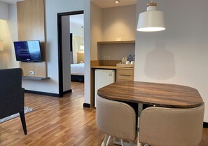 Moderni hotellihuone, jossa on puinen ruokapöytä, kaksi tuolia, seinälle asennettu televisio, keittiö sekä ovi, joka johtaa toiseen huoneeseen.