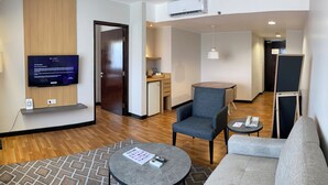 Habitación de hotel moderna con televisor de pantalla plana, sofá, sillón, una mesita y una kitchenette al fondo.