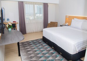 A hotel room with a bed, a desk, a chair, a television, and a window with curtains. - Grand Dafam Braga Bandung (Bandung)