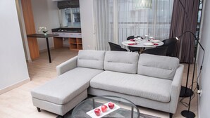 Una sala moderna con un sofá seccional gris, una mesa de centro de vidrio con manzanas, y un comedor al fondo.