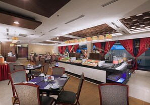A buffet-style restaurant with multiple dining tables and chairs, a central serving counter with food displays, and a modern ceiling design. - Grand Dafam Braga Bandung (Bandung)