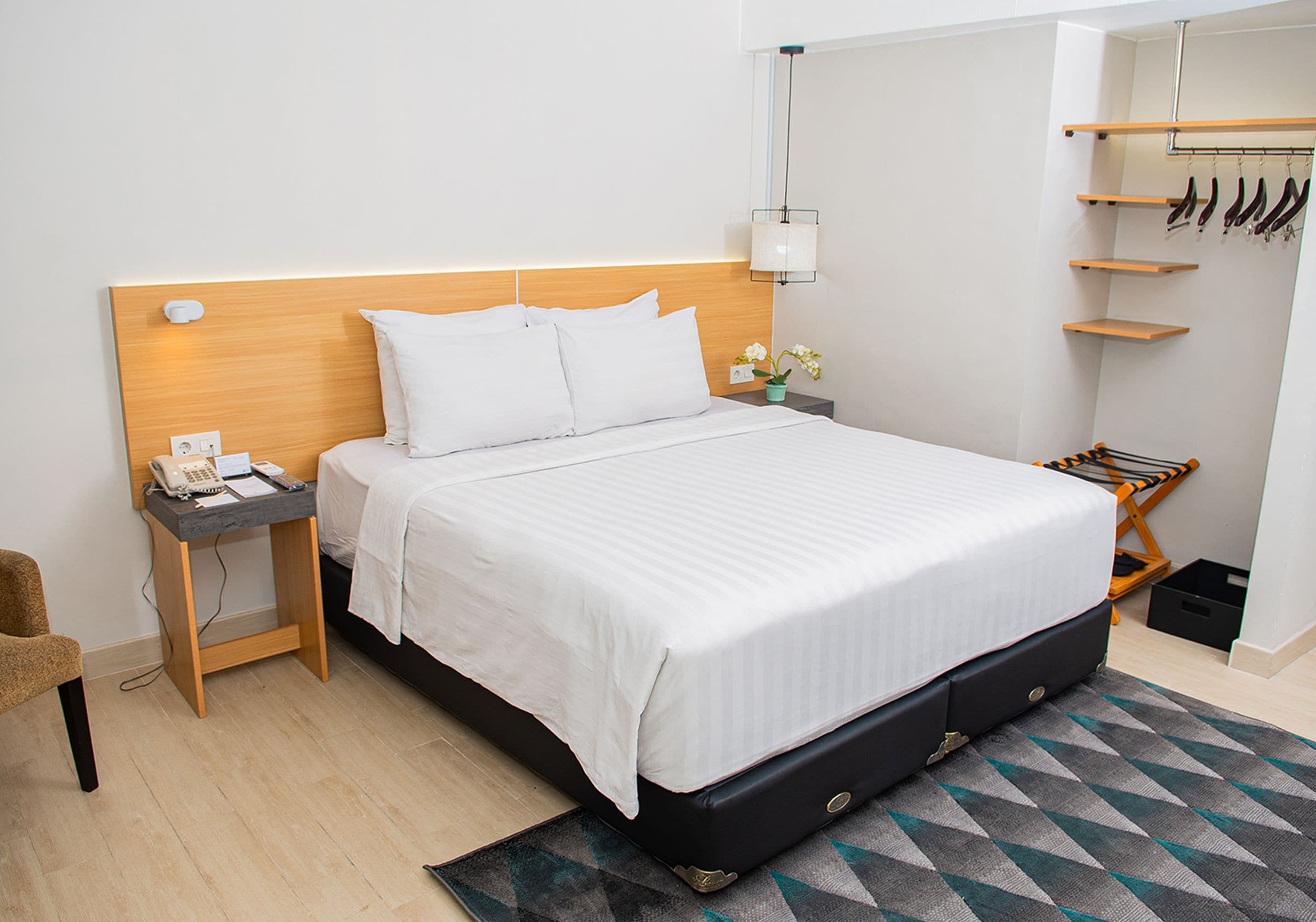 A neatly arranged hotel room with a large bed, a bedside table with a phone and notepad, a small nightstand with a lamp, and a shoe rack against the wall.