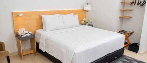 Habitación de hotel ordenada con una cama grande, una mesita de noche con teléfono y bloc de notas, una mesita auxiliar pequeña con lámpara y un zapatero contra la pared.