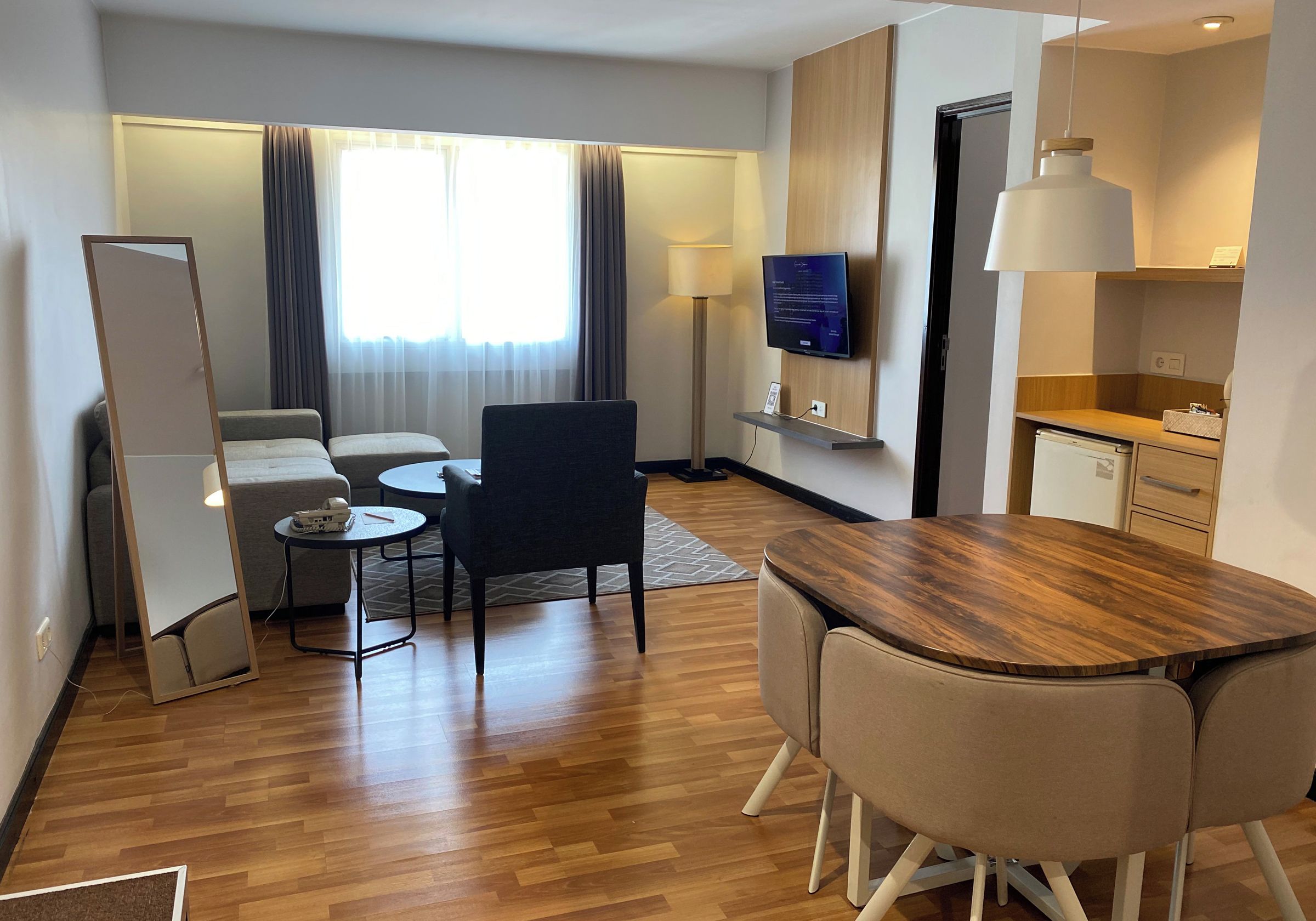 A hotel room with a wooden floor, a sofa, a dining table with chairs, a TV mounted on the wall, and a small kitchen area.