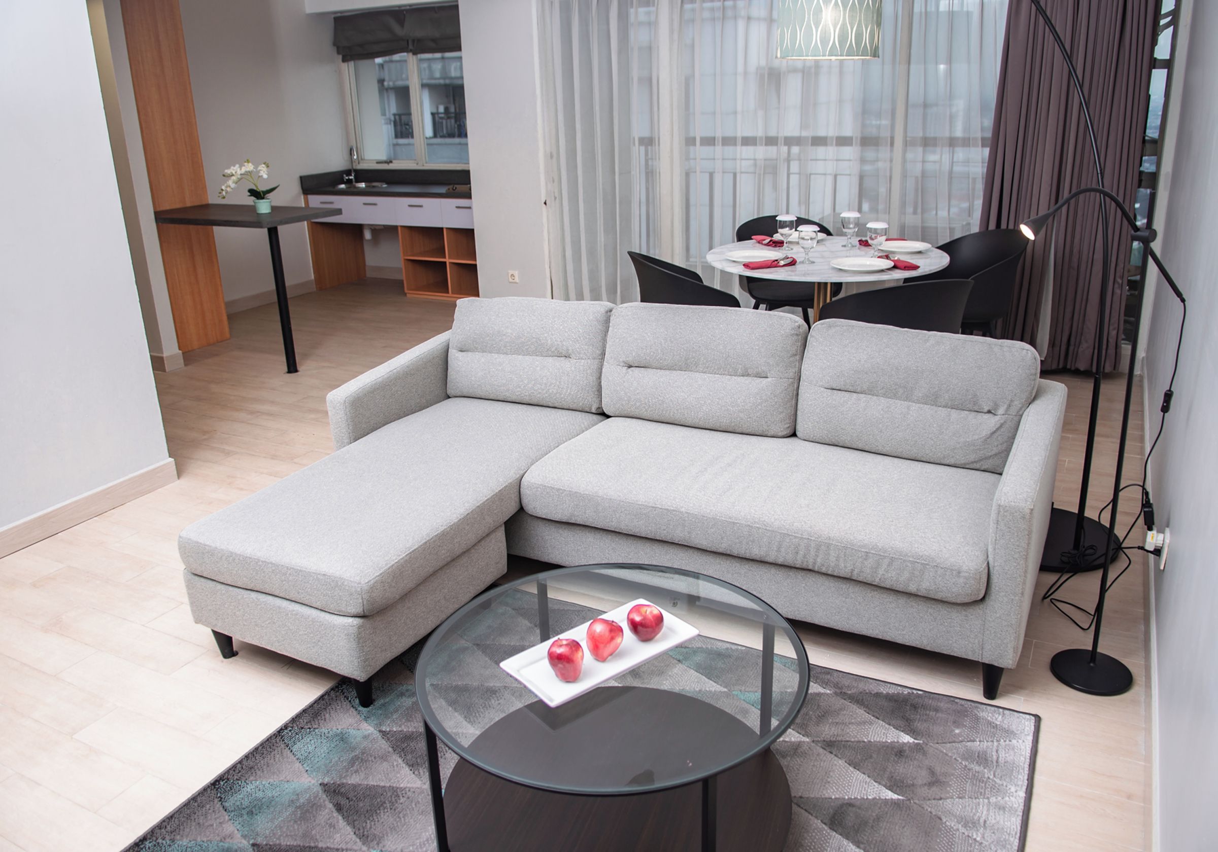 A modern living room with a grey sectional sofa, a glass coffee table with apples, and a dining area in the background.