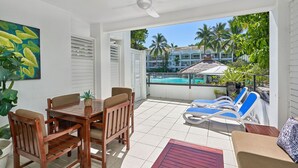 A balcony with a wooden table and chairs, a pool in the background, and palm trees. - Beach Club Lagoon View 2 Bedroom Apartment 3113-4 (Palm Cove)