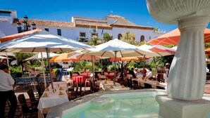 Un área de comedor al aire libre con mesas y sombrillas, una fuente y un edificio al fondo.