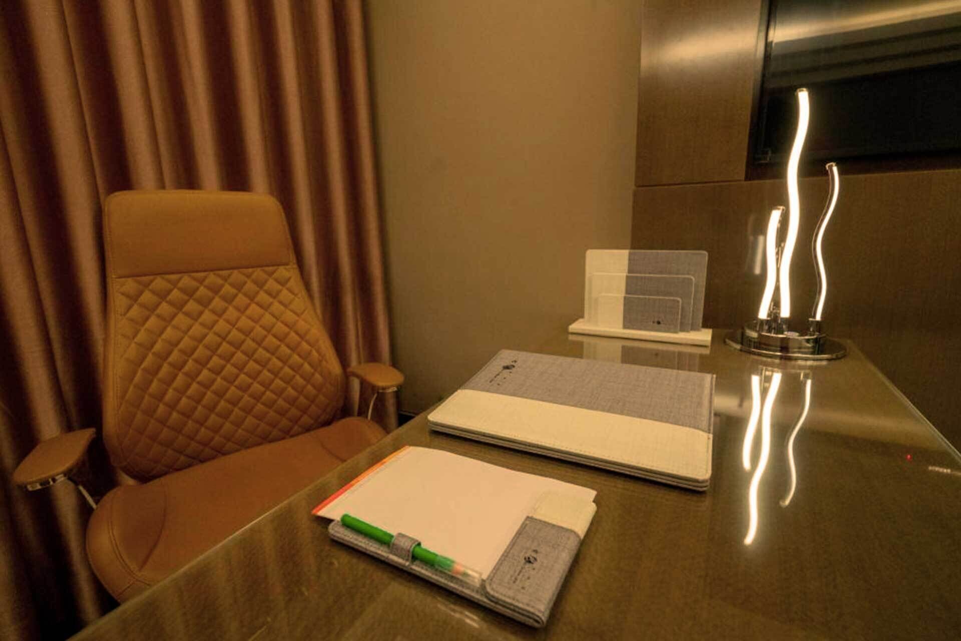 A hotel room with a leather chair, a desk with a notebook, a pen, a mobile phone, and a lamp.