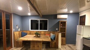 Habitació amb taula de dinar de fusta, una banca integrada, una planta petita, una finestra, mobles prefabricats per a llibres i un frigorífic.