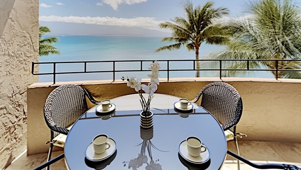 Een ronde tafel met twee stoelen, een vaas met witte bloemen, en twee kopjes op een balkon met uitzicht op een strand met palmbomen en de oceaan.
