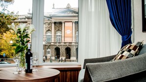 Una habitación con vista a un edificio histórico, una mesa con una botella y vasos, y un sofá con un cojín a cuadros.