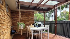 Une terrasse couverte avec une plateforme en bois, une table et des chaises, des plantes suspendues et un barbecue.