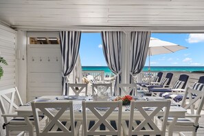A covered outdoor dining area with white furniture, striped curtains, and a view of the ocean.