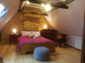 Un dormitorio acogedor en el ático, con techo inclinado, cama de madera, mesita de noche, cómoda y un pequeño otoman.