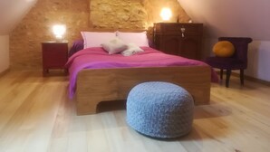 Un dormitorio con techo inclinado, una cama de madera, una mesita de noche, un aparador, una silla y un otoman redondo.