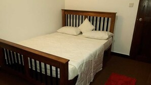 Una cama individual con estructura de madera y ropa blanca, una alfombra roja en el suelo y una puerta cerrada al fondo.