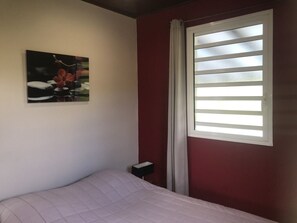 Une chambre à coucher comprenant un lit, une table de chevet, une fenêtre avec des stores et un tableau accroché au mur.