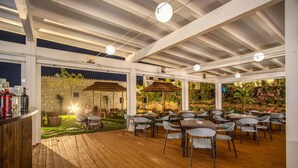 An outdoor patio with wooden flooring, tables, and chairs, surrounded by a stone wall and greenery. - Cala Sinzias Resort (Castiadas)