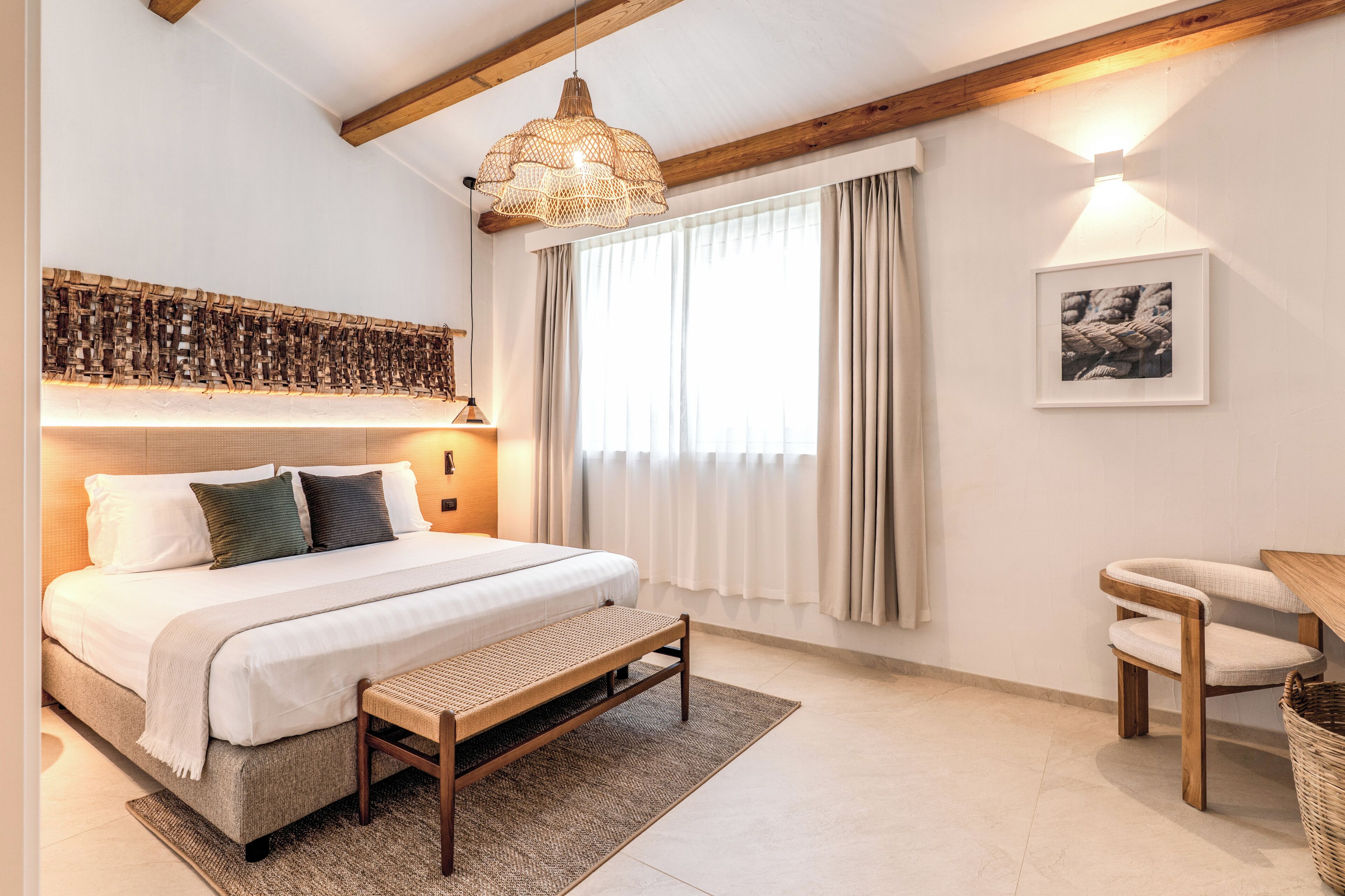 Een slaapkamer met een bed, een bankje, een stoel, een hanglamp, een aan de muur bevestigd schilderij en een raam met gordijnen.