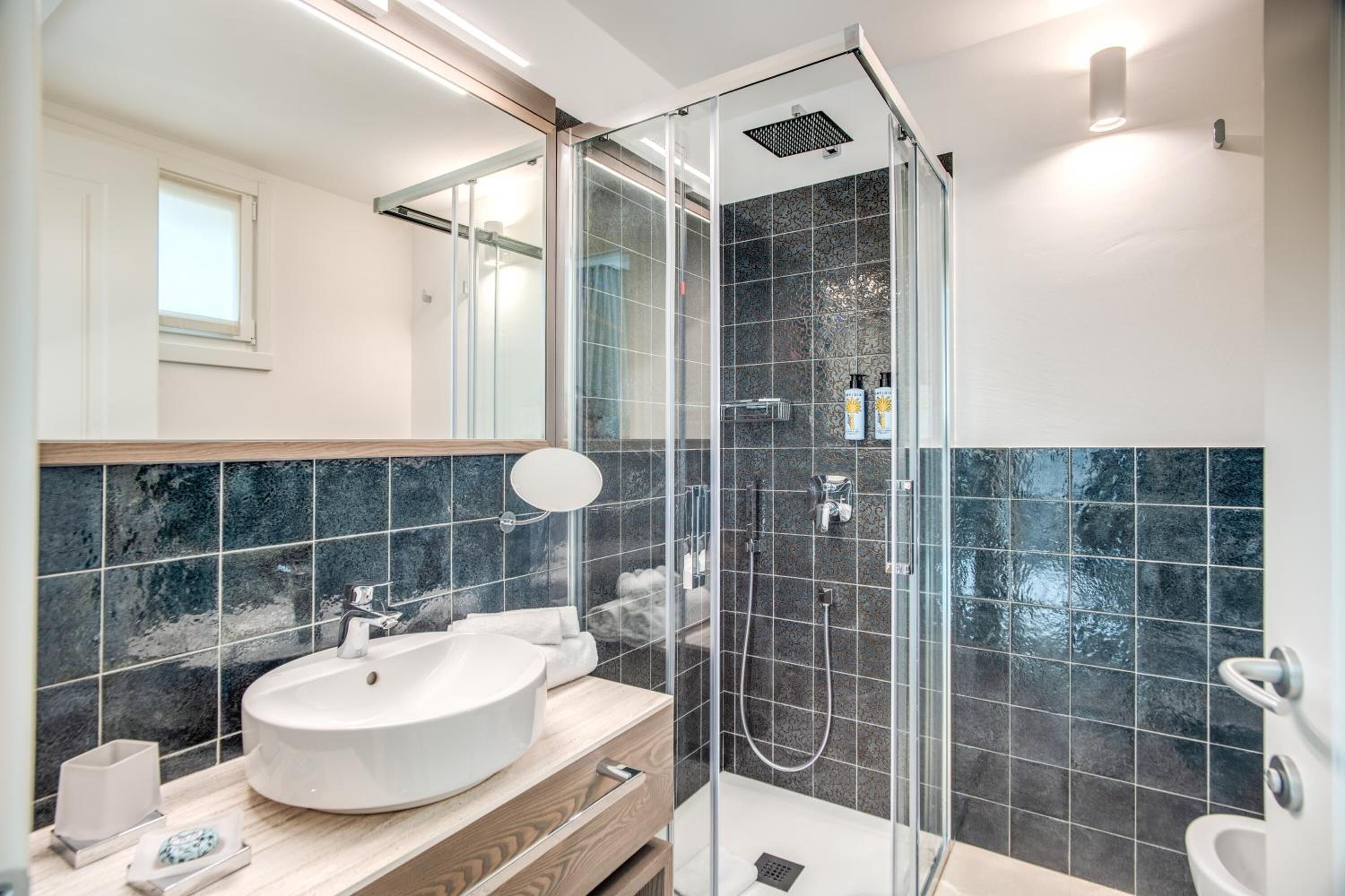 Een moderne badkamer met een groot wit wastafel, een douchecabine van glas en donkere betegelde wanden.