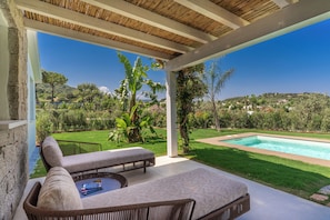 A covered outdoor area with a pool, lounge chairs, and a view of a lush garden and hills. - Cala Sinzias Resort (Castiadas)