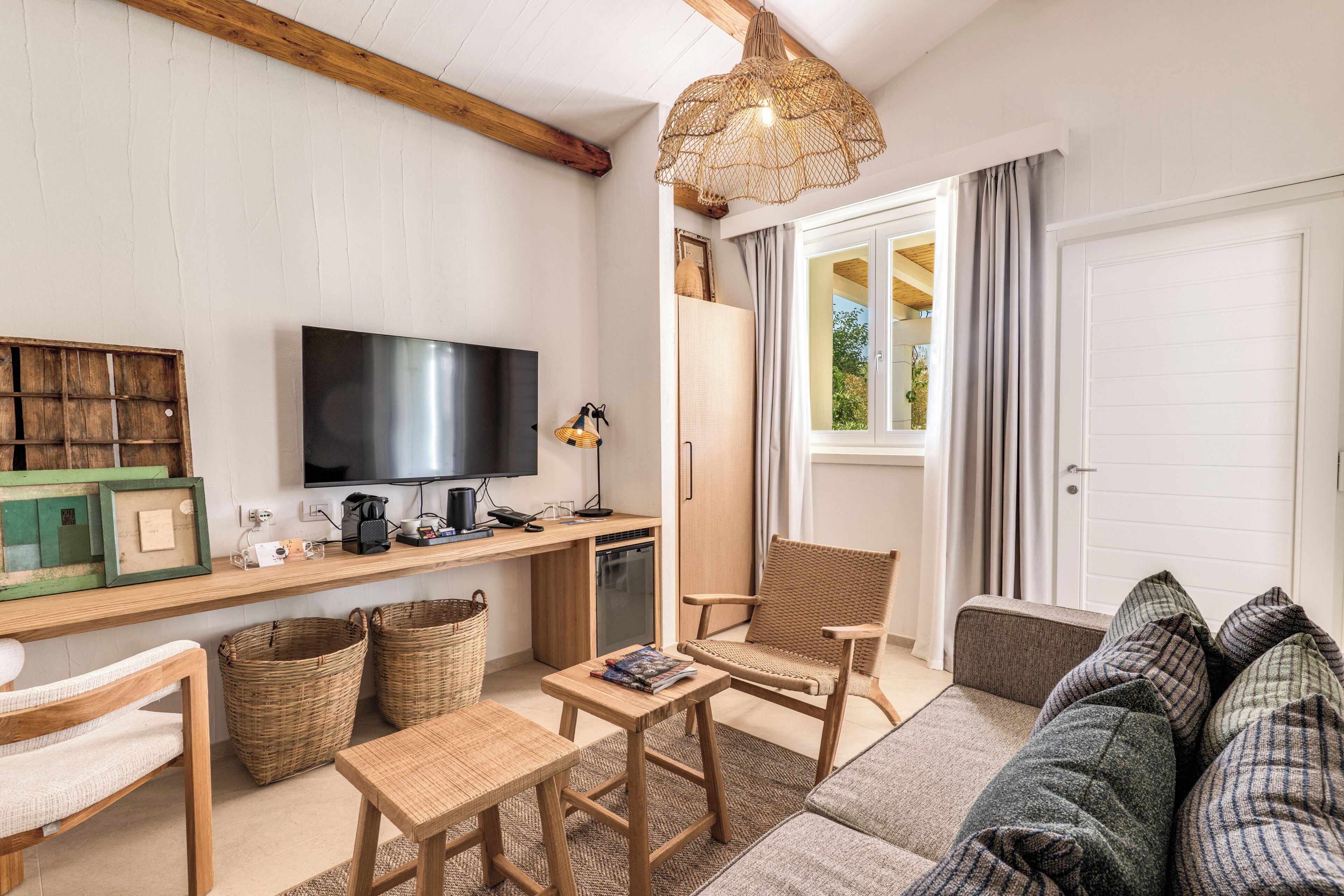Ein möbliertes Wohnzimmer mit Fernseher, Holzmöbeln, Korbwannen und einem großen Fenster mit Vorhängen.