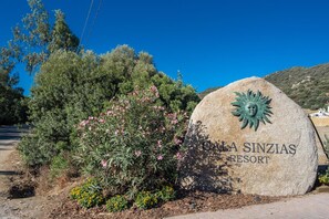 A large stone sign with the text "LA SINZIAS RESORT" and a sun emblem, surrounded by greenery and flowers. - Cala Sinzias Resort (Castiadas)
