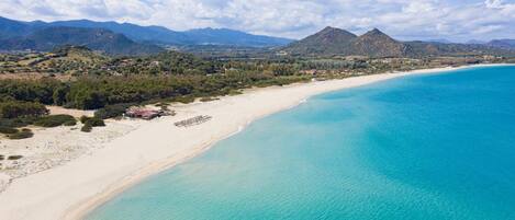 A coastal landscape with a sandy beach, clear turquoise waters, and distant mountains under a partly cloudy sky.
