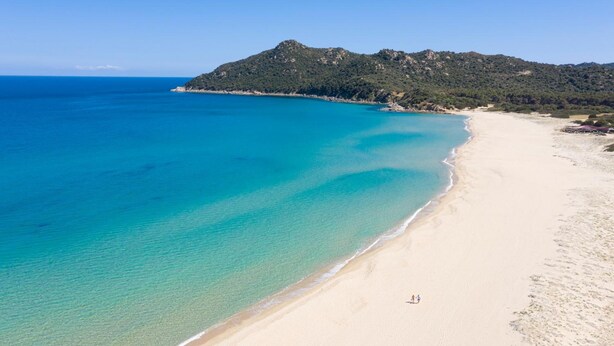 A beach with clear turquoise waters, a sandy shore, and a forested hill in the background. - Cala Sinzias Resort (Castiadas)