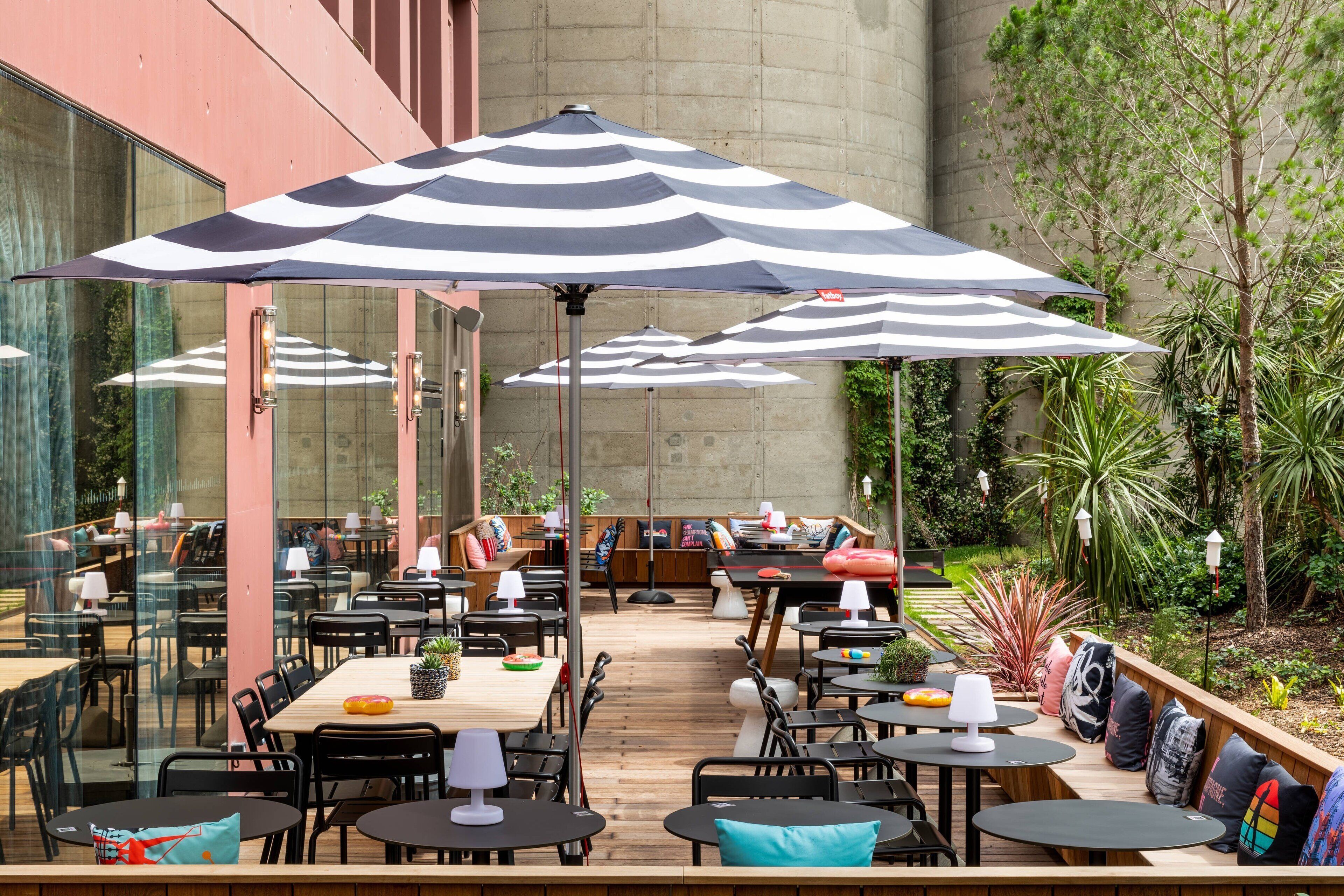 Een terras met meerdere tafels en stoelen, grote gestreepte parasols, en een tuin met planten en bomen.