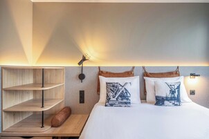 A hotel room with a bed, two pillows with a cityscape design, a wooden nightstand, a lamp, and a shelf unit.