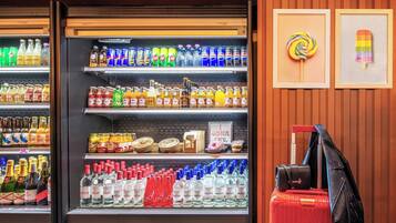 A beverage refrigerator stocked with various drinks, a luggage trolley, and two framed artworks on the wall.