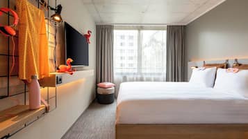 Una habitación de hotel moderna con una cama grande, un televisor montado en la pared, una ventana con cortinas y una mesita redonda con un cojín rosa.
