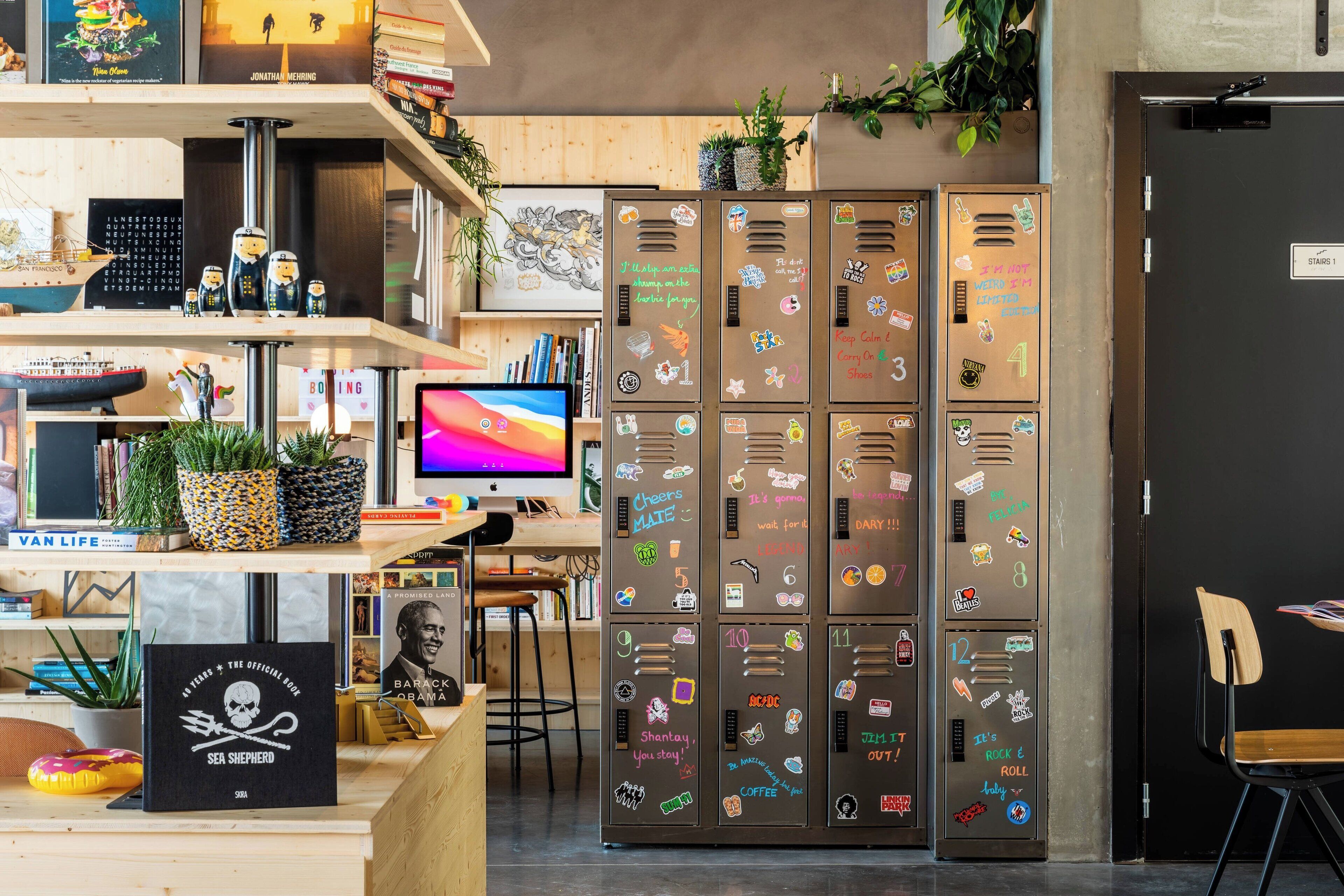 Ein moderner Arbeitsraum mit Schreibtisch, Computer, Regalen mit Büchern und Dekoration sowie Schließfächern mit verschiedenen Aufklebern.