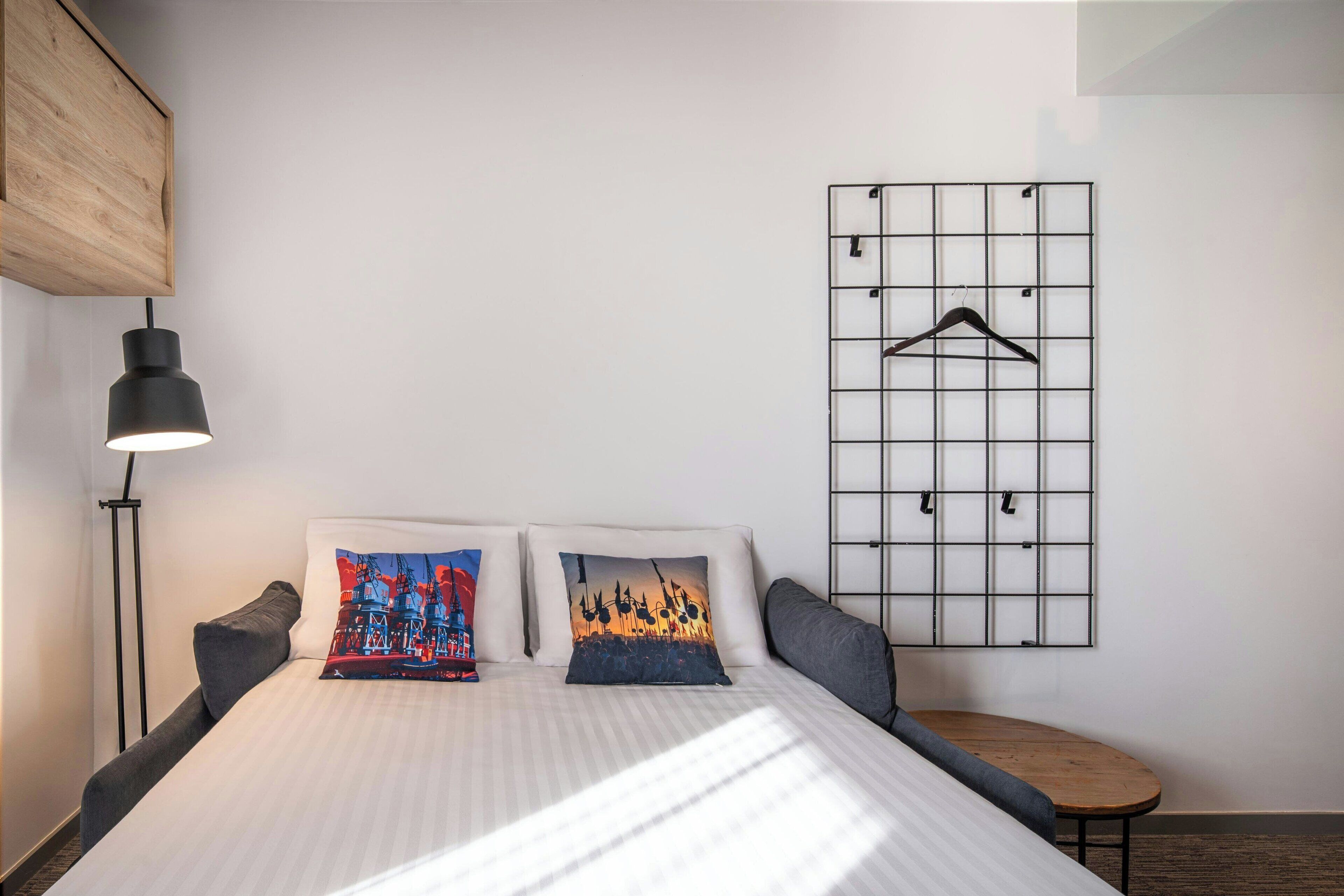 Une chambre d’hôtel bien aménagée, avec un lit, des oreillers, un luminaire suspendu, une petite table et un panneau mural avec un porte-manteau.